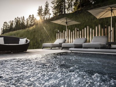 Il nostro hotel con jacuzzi in Alto Adige Vasca idromassaggio con lettini e ombrelloni davanti a pendio boscoso al tramonto