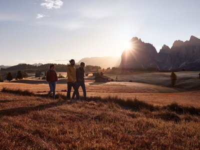 Hotel Schmung: hiking holiday at Alpe di Siusi/Seiser Alm Three hikers at sunset on a mountain meadow with a cabin in the background