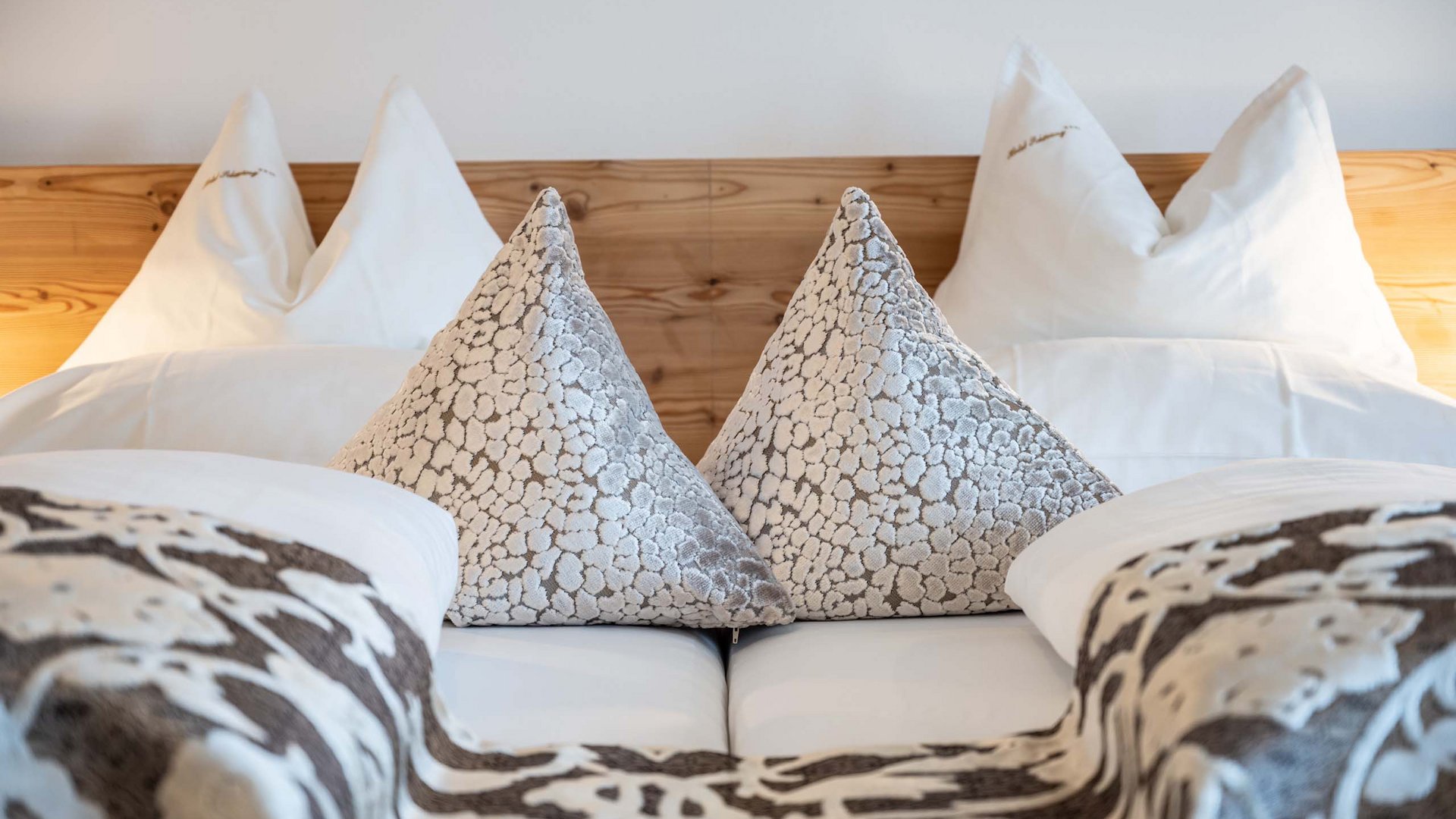 I nostri servizi inclusi per la vostra vacanza in Alto Adige Letto matrimoniale con due cuscini decorativi e coperte a motivo in cornice di legno chiaro